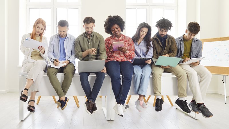 Junge Menschen die nebeneinander auf einem Tisch sitzen und in Laptops und Tablet schauen.